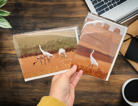 Two greeting cards with Whooping Crane designs on a wooden desk with a laptop and coffee cup.