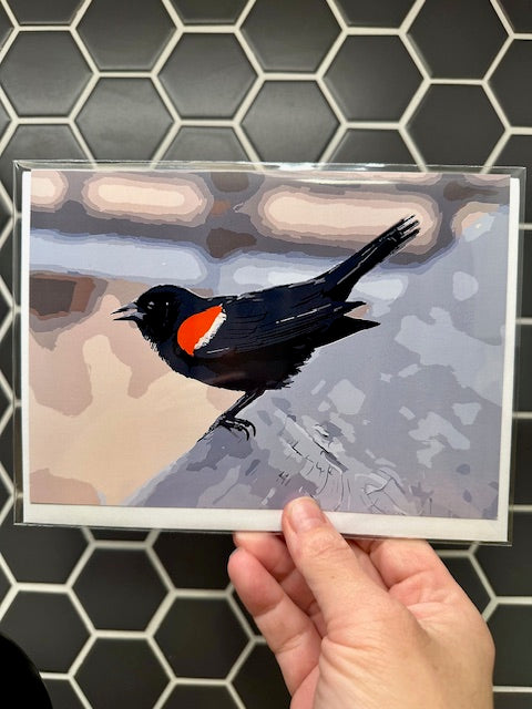 Hand holding a card with a Red-winged Blackbird illustration against a tiled wall background